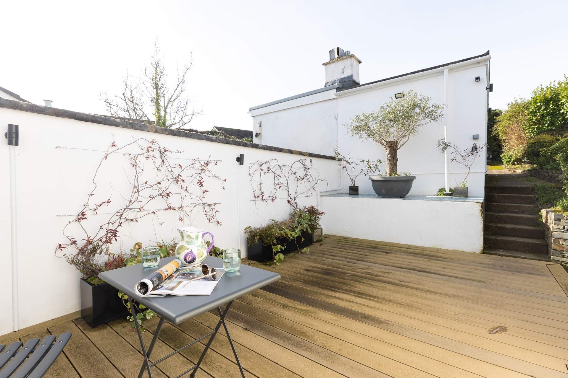 The rear decked area has a Mediterranean vibe with white washed walls and bistro table. The steps lead to the parking area and garage with washing machine and tumble-drier.